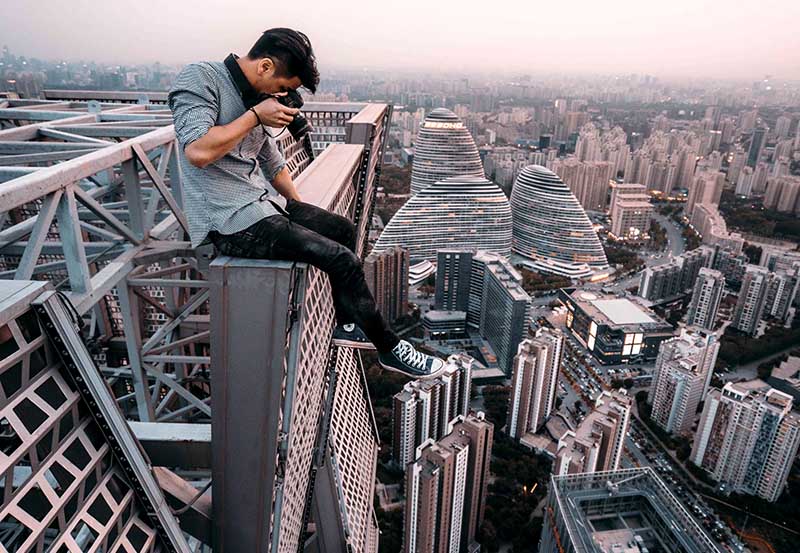 a young man casually checks his camera while sitting on the edge of a skyscraper